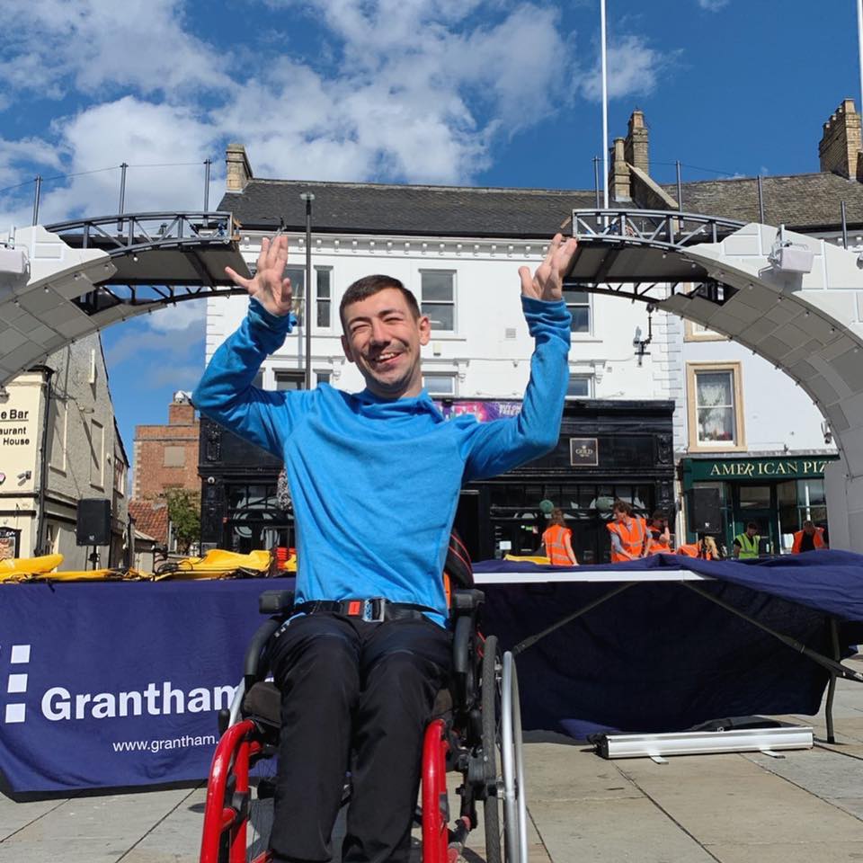 A man in a wheelchair sits in front of the broken bridge. He is raising his hands into the air, and seems to be holding each side of the top of the broken bridge.