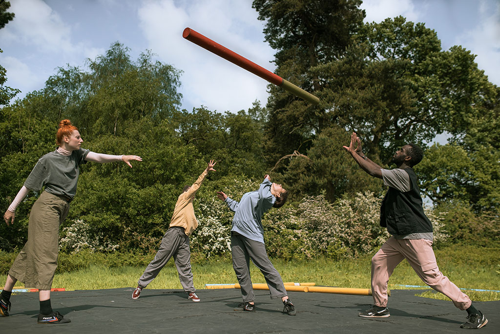 The four dancers throw a heavy wooden pole into the air. Each person is captured mid-movement, with arms raised and legs stretched.