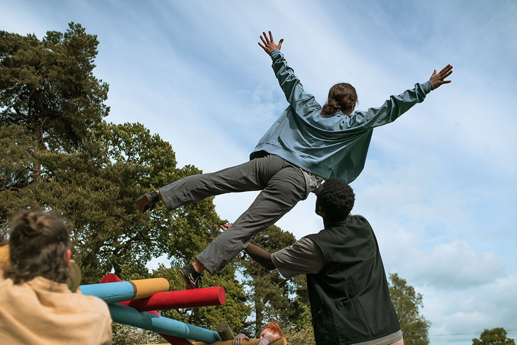 A dancer stands on top of the free-standing bridge, her hands stretched into the sky. She balances on one foot whilst another dancer supports her.