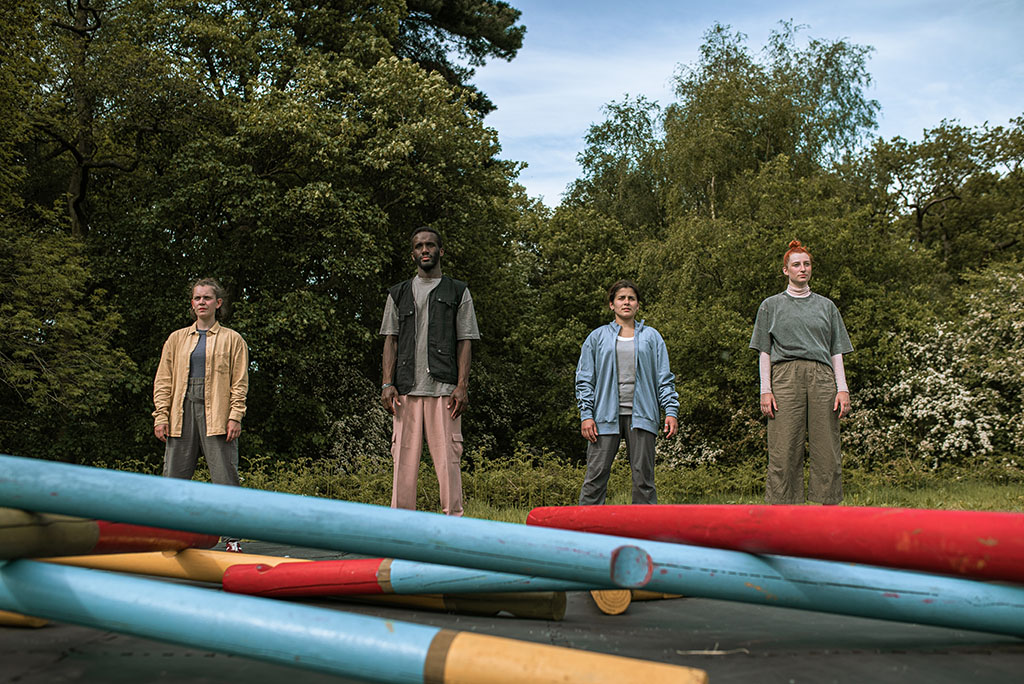 Four dancers face the viewer in a line. In front of them are logs that will form the basis of a free-standing self-supporting bridge. The dancers wear loose, comfortable clothing in plain colours.