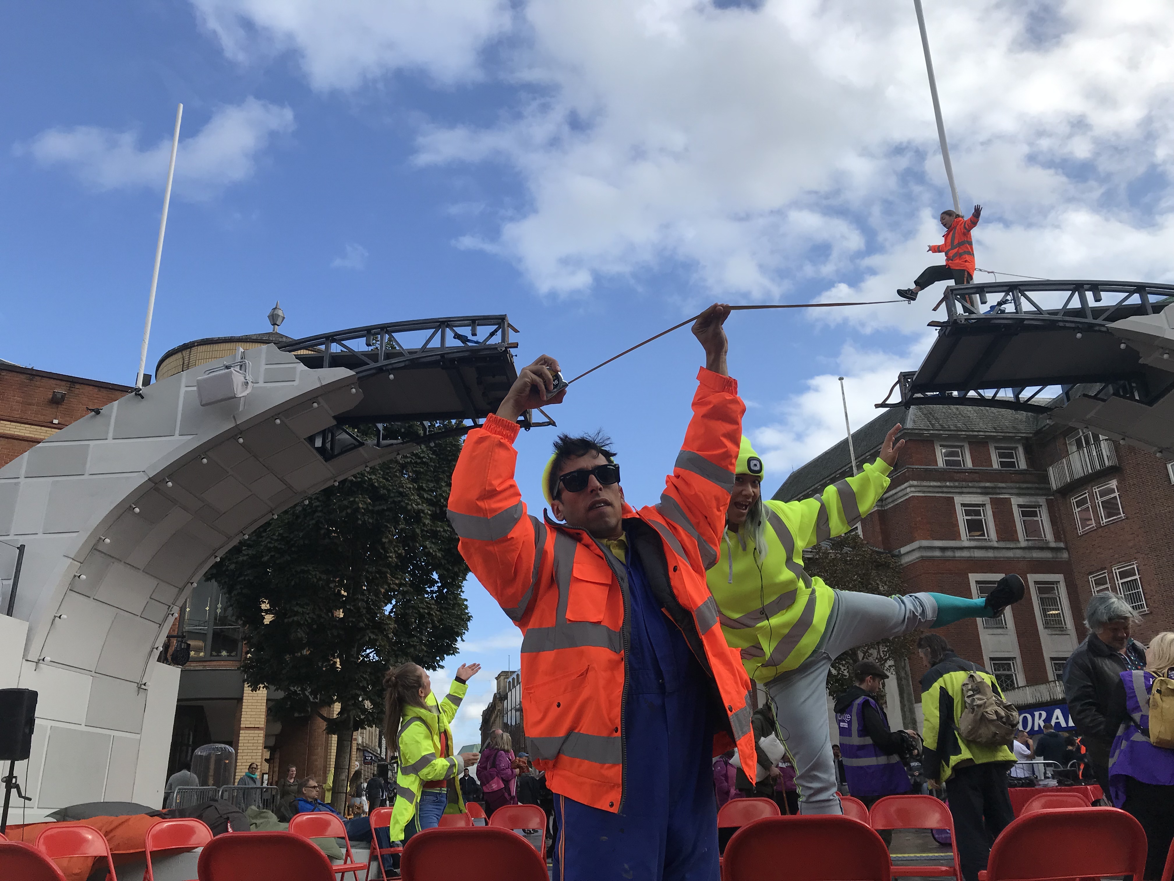 Two clowns wearing high vis clothes hold out a tape measure against the bridge. Up high, a performer steps out and appears to be walking on the tape measure. 