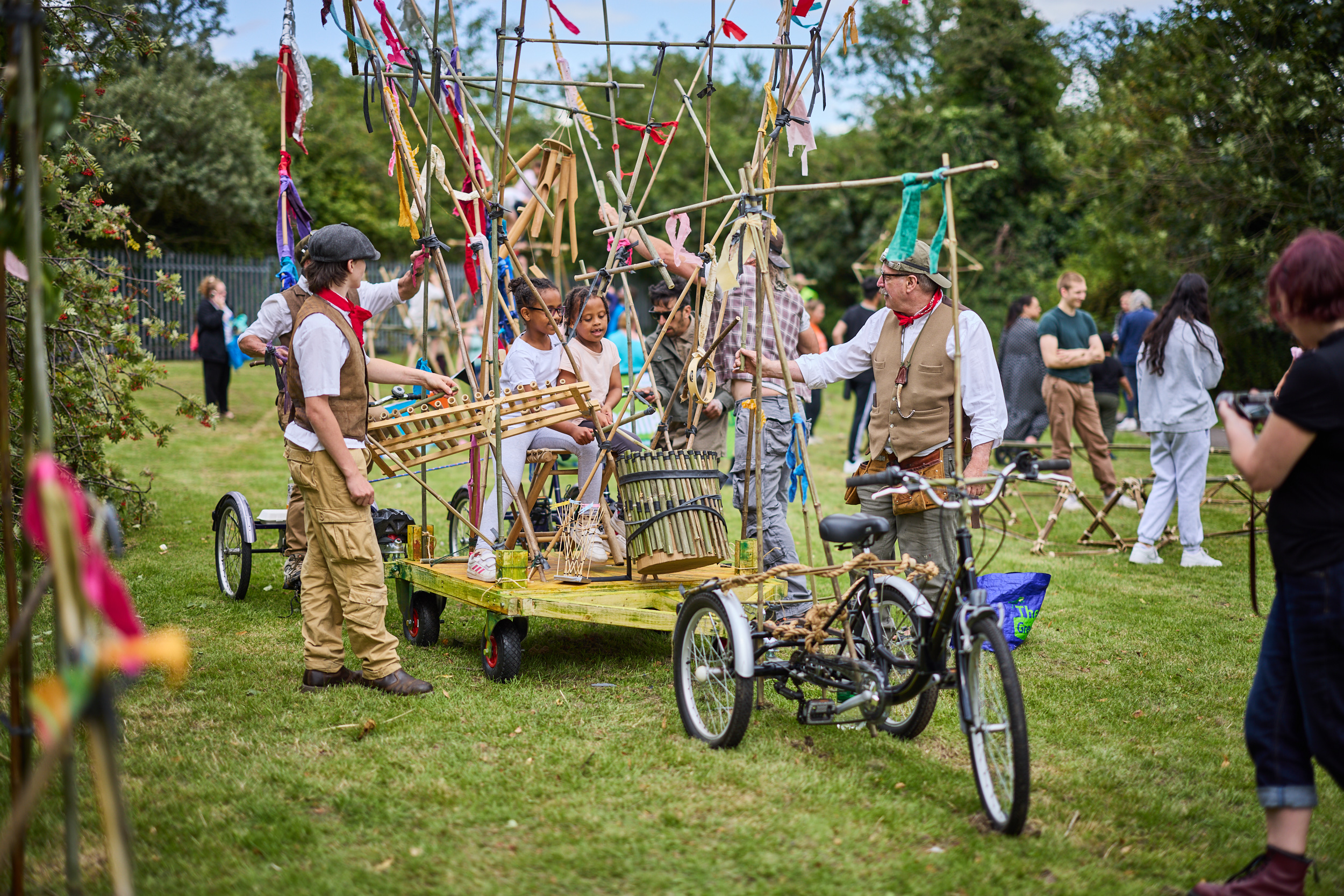 A trike holds a trailer filled with  bamboo structures, flags, musical instruments and children