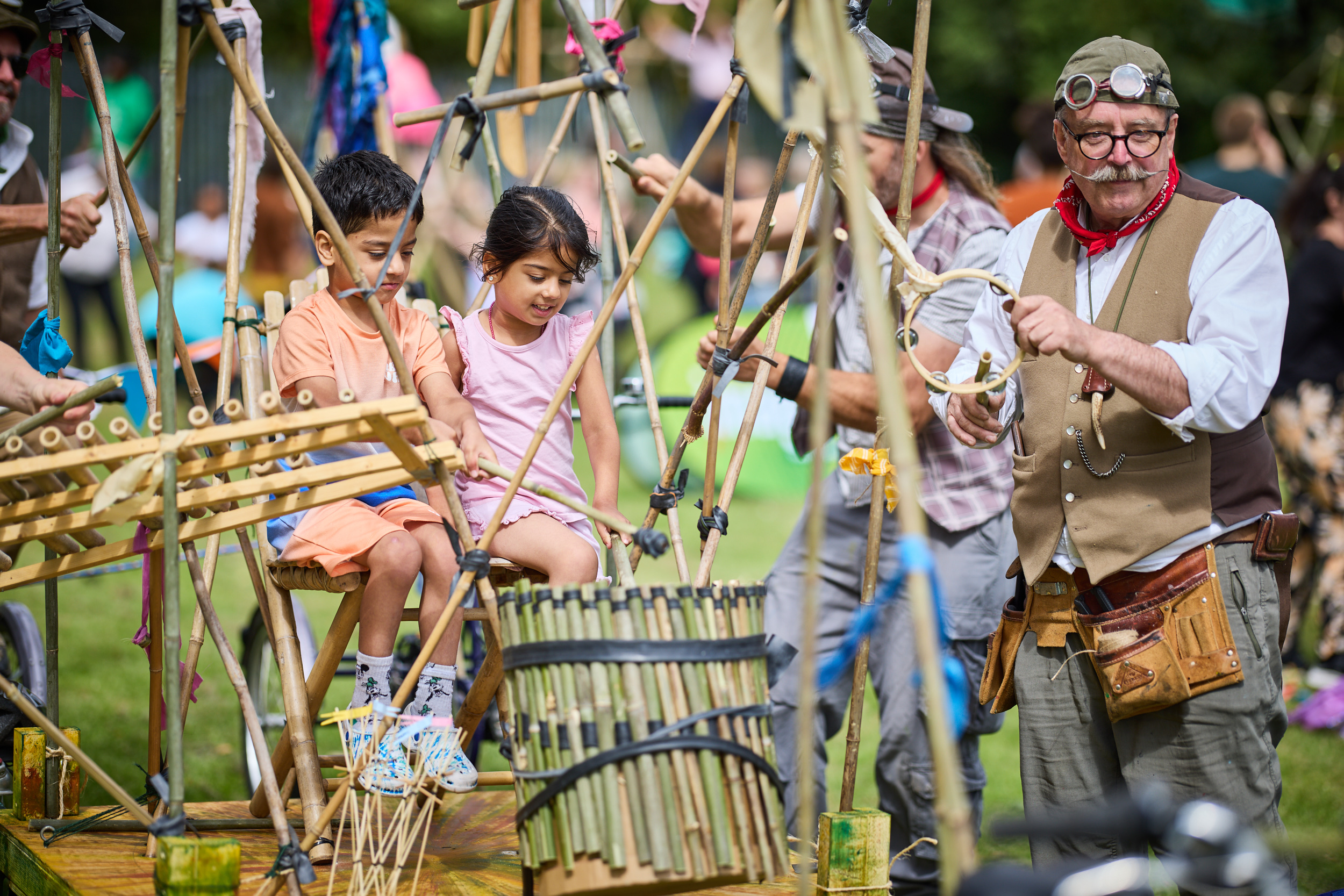 Two children sit in a contraption made of bamboo, with a xylophone and tambourine