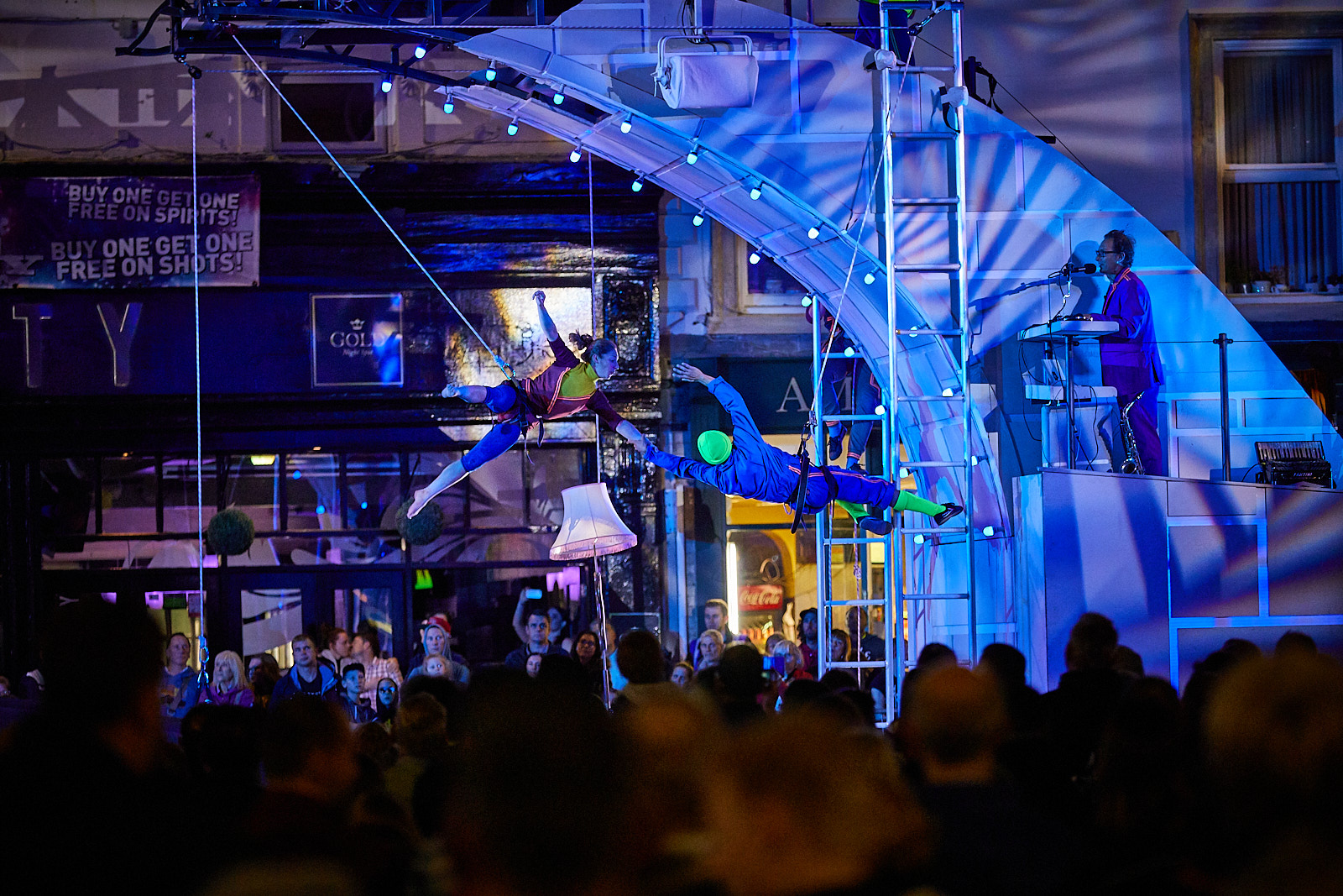 A nighttime shot of the Bridge performance. A trapeze artist hangs from a wire, where she is caught by a man handing vertically from a ladder suspended from the bridge.