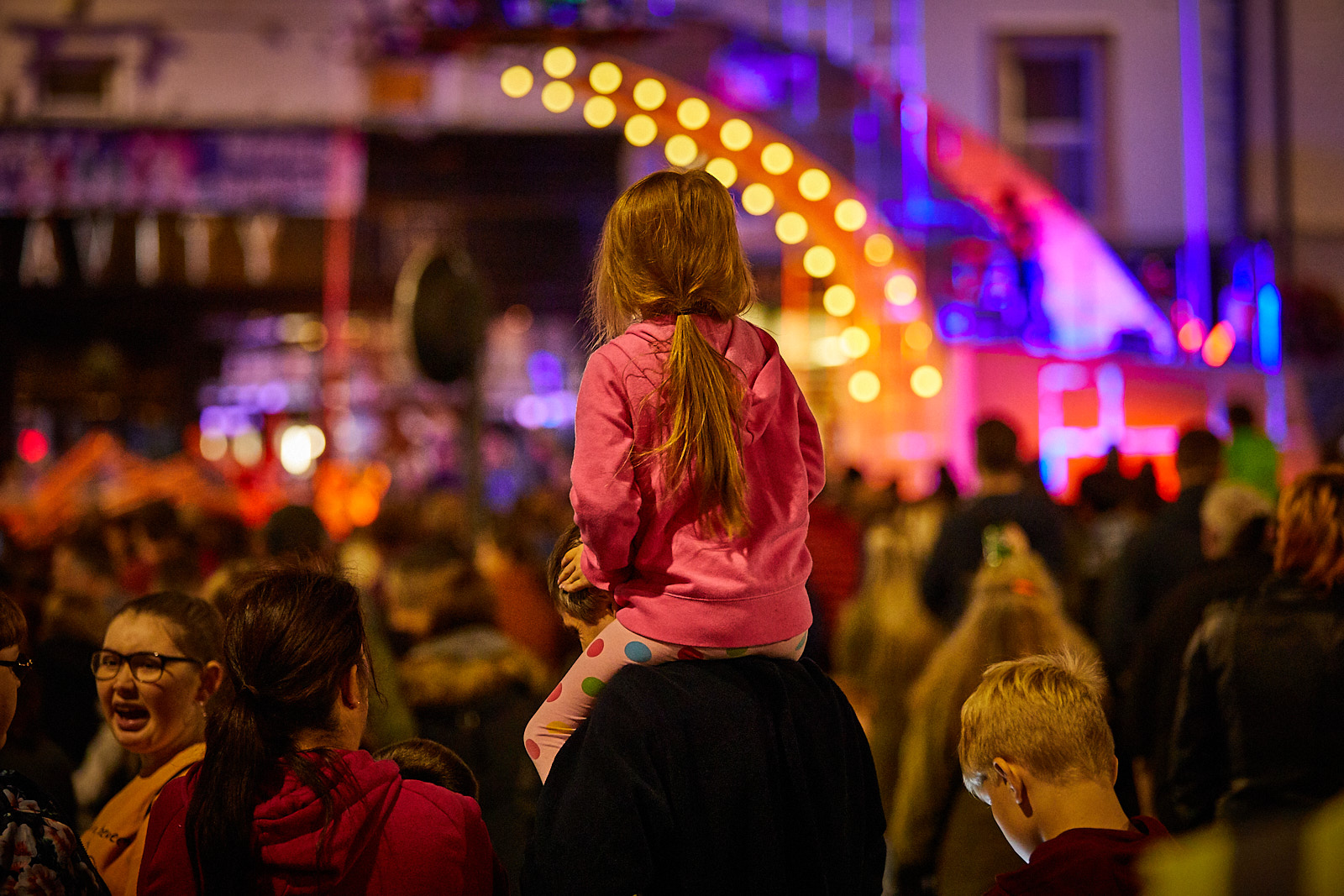 A young girl sits on her dad's shoulders, looking over the crowd as a performance begins. The bridge is colourfully lit.