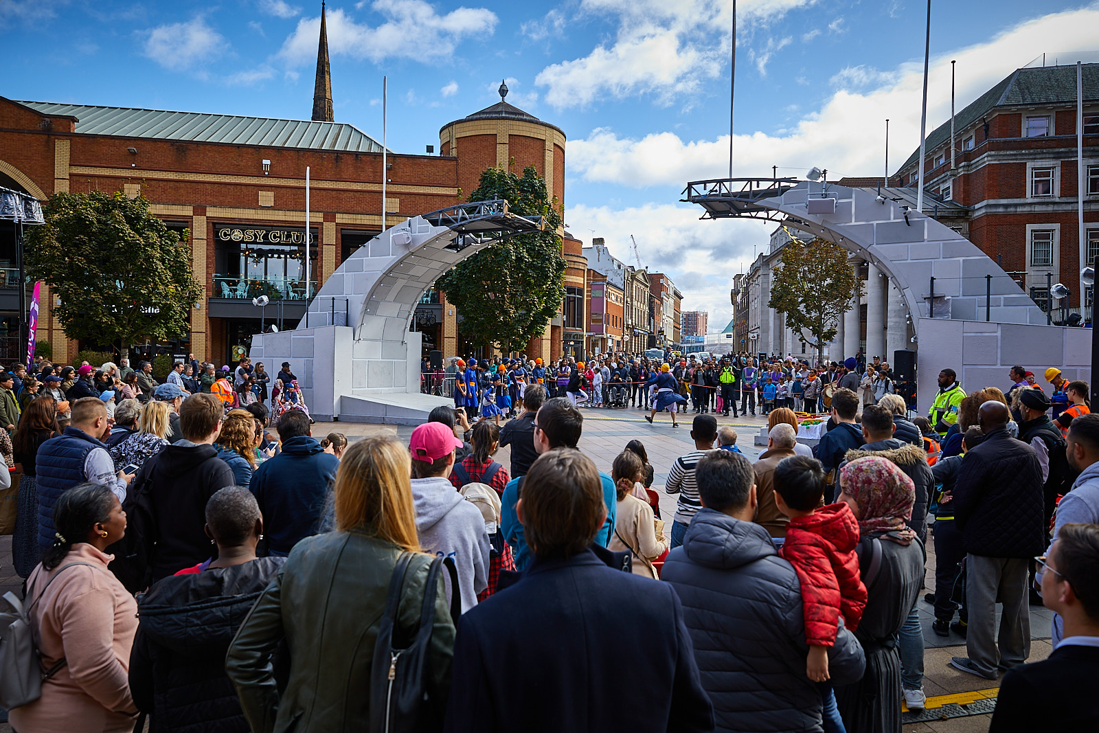 A broken bridge is in a busy city-centre square. It is surrounded by a crowd of onlookers. Below the bridge, Sikh dancers perform in blue costume with orange turbans.