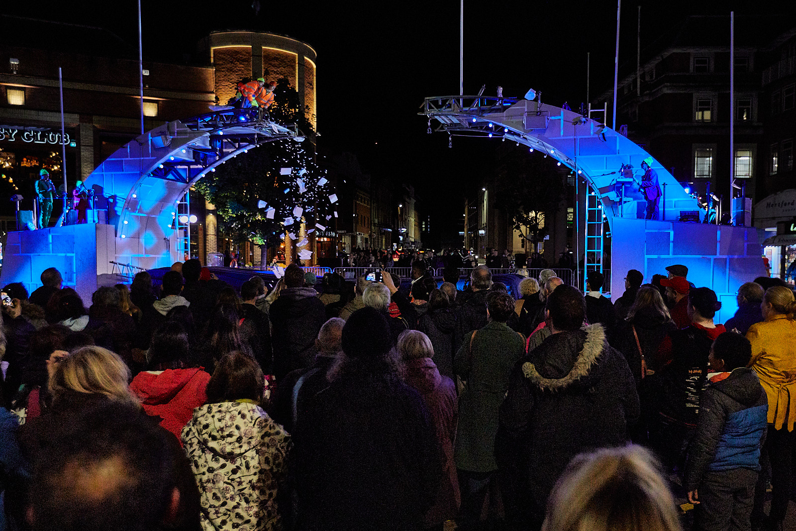 It is nighttime and the broken bridge is dramatically lit in blue. Actors are at the top of the bridge, looking down as a shower of paper scatters to the ground. A large audience looks on at the performance.