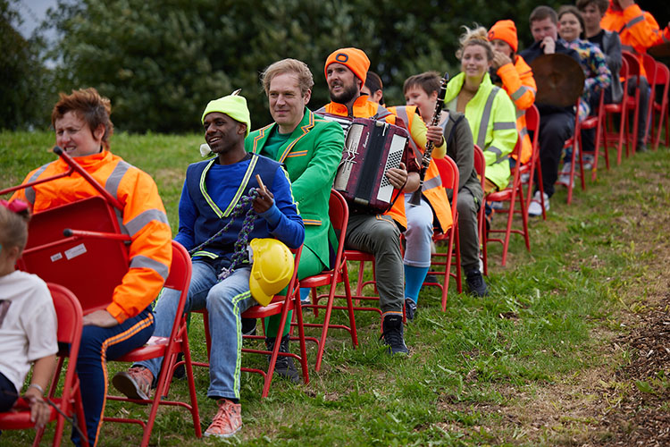 Performers with instruments and bright high-vis costumer sit on red chairs positioned in a line, facing down the hill on grass
