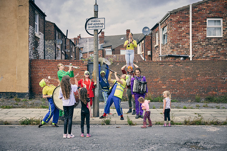 Musicians in high-vis costume stand on the street surrounded by children. Behind them are terraced houses. An old tyre balances from a road sign.