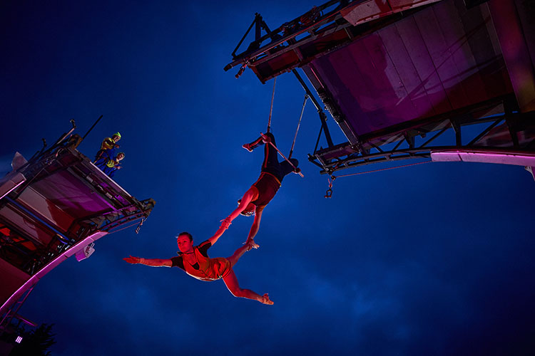 Two acrobats perform, one hangs from a swing on the bridge, she holds her partner's arm and leg as they perform in mid-air