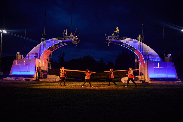 Four local people carry logs as part of the performance. Behind them the broken bridge is lit against the night sky.