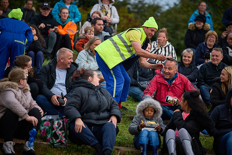 A clown in high vis costumer entertains the crowd by combing the hair of an audience member