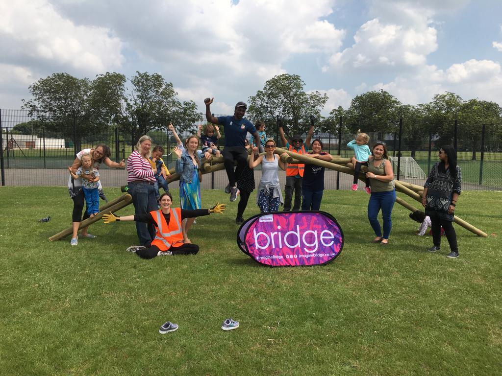 A group of adults and children are perched in and around the self-supporting bridge, all in celebration at their efforts. A pop-up sign that reads BRIDGE is infront of the bridge.