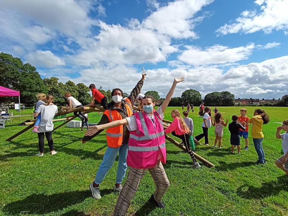 Two people stand in front of the self-supporting bridge. They are striking a pose, hands in the air and hips jutting to one side. Behind them are children and families having fun around the bridge.